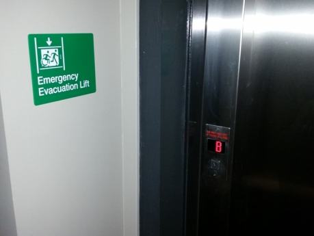 Emergency Evacuation Lift sign on a wall next to the lift doors, in green, with Braille and Tactile characters, and accessible means of egress icon wheelchair symbol-min.jpg