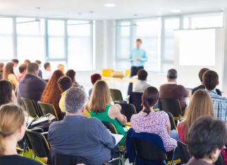 A view from the back of a corporate training session, with people seated looking towards a person standing next to a screen
