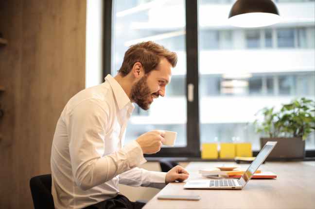 man holding teacup in front of laptop on top of table inside the room