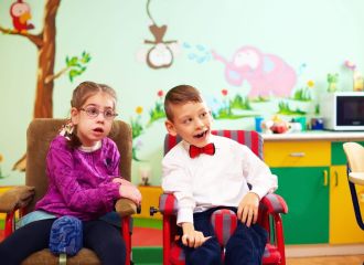 Two young kids, a boy and a girl, both with disabilities, sitting in a colourful classroom